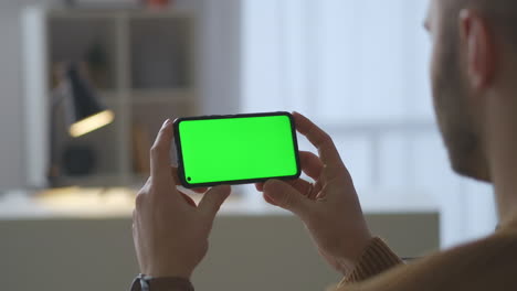 Greenscreen-Auf-Modernem-Smartphone-In-Den-Händen-Eines-Mannes,-Der-Zu-Hause-Sitzt-Und-Nahaufnahme-Eines-Videos-Durch-Die-Schulter-Eines-Männlichen-Benutzers-Ansieht